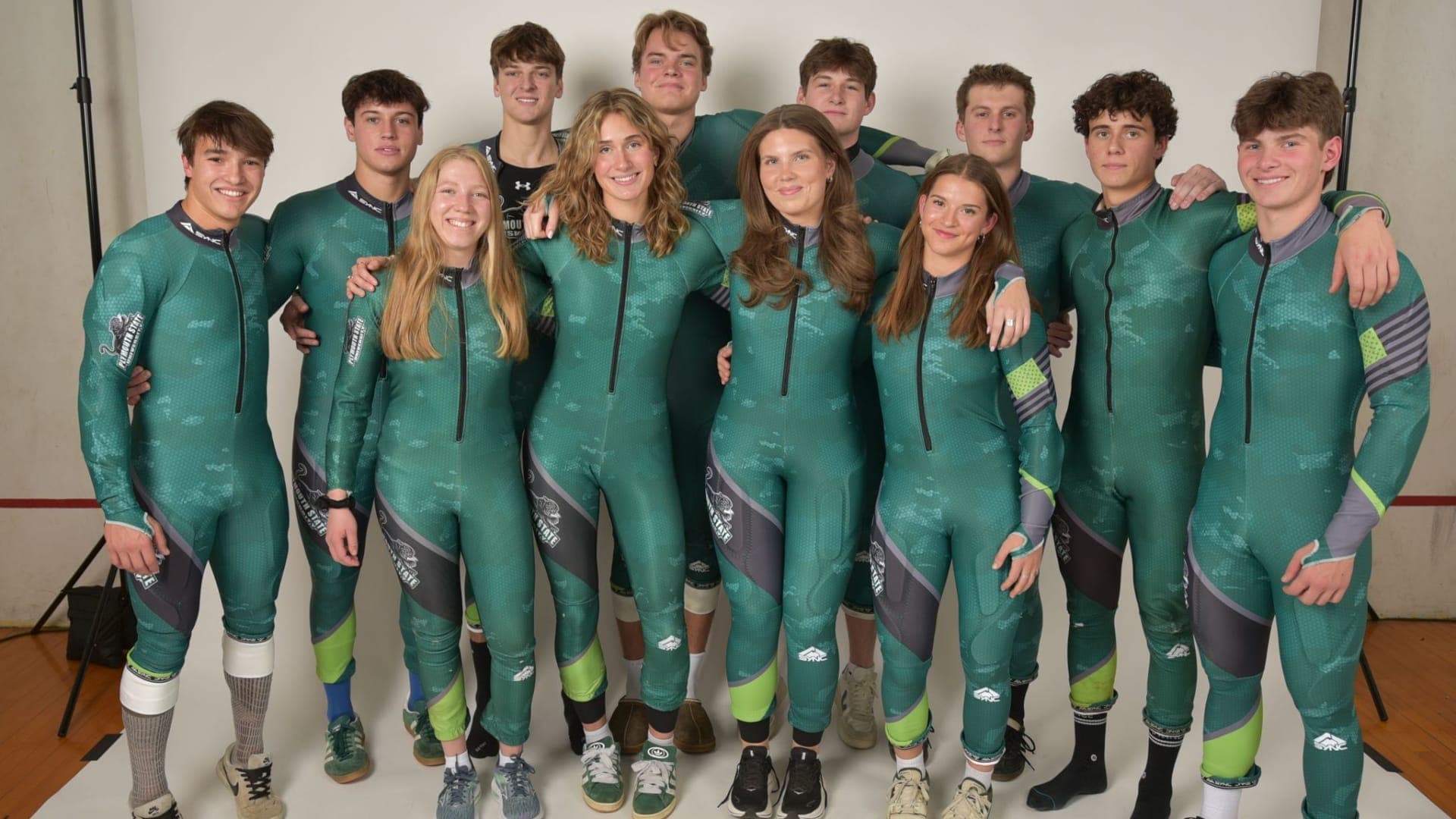 Plymouth State University Ski Team | Julia Müller Plymouth State University ski team in front of a white wall in green skiing racing suits.