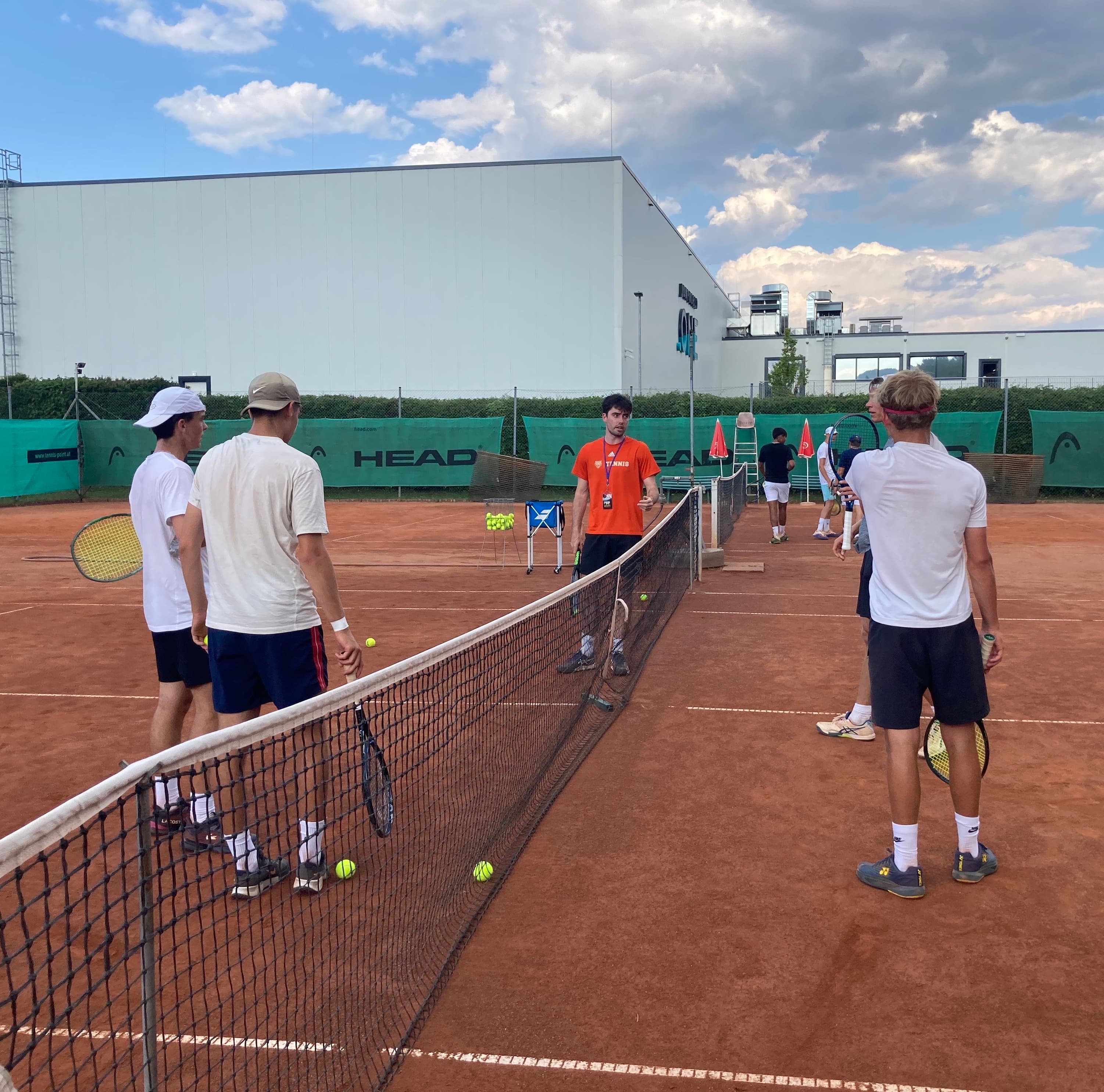 Tennis Practice Session with U.S. College Coach at the Smarthlete Showcase A group of tennis players standing on a red clay court during a practice session, listening to a U.S. college coach in an orange shirt.